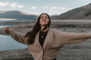 A woman standing on rocky terrain near water, arms extended, symbolizing healing and renewal.