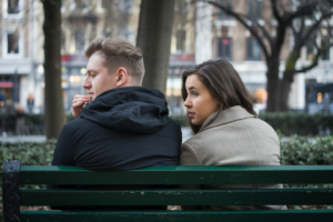 A couple sitting on a bench, one partner looking thoughtful and the other avoiding eye contact, symbolizing conflict avoidance in relationships.