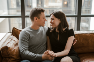 A couple sitting on a sofa in a modern living room, holding hands and looking at each other.